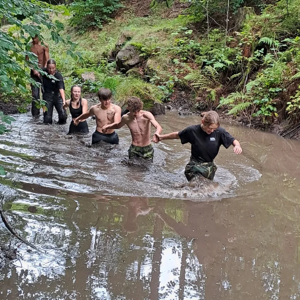 Studenti v maskáčových kalhotách brodí bahnitým potokem v lese během branného kurzu.