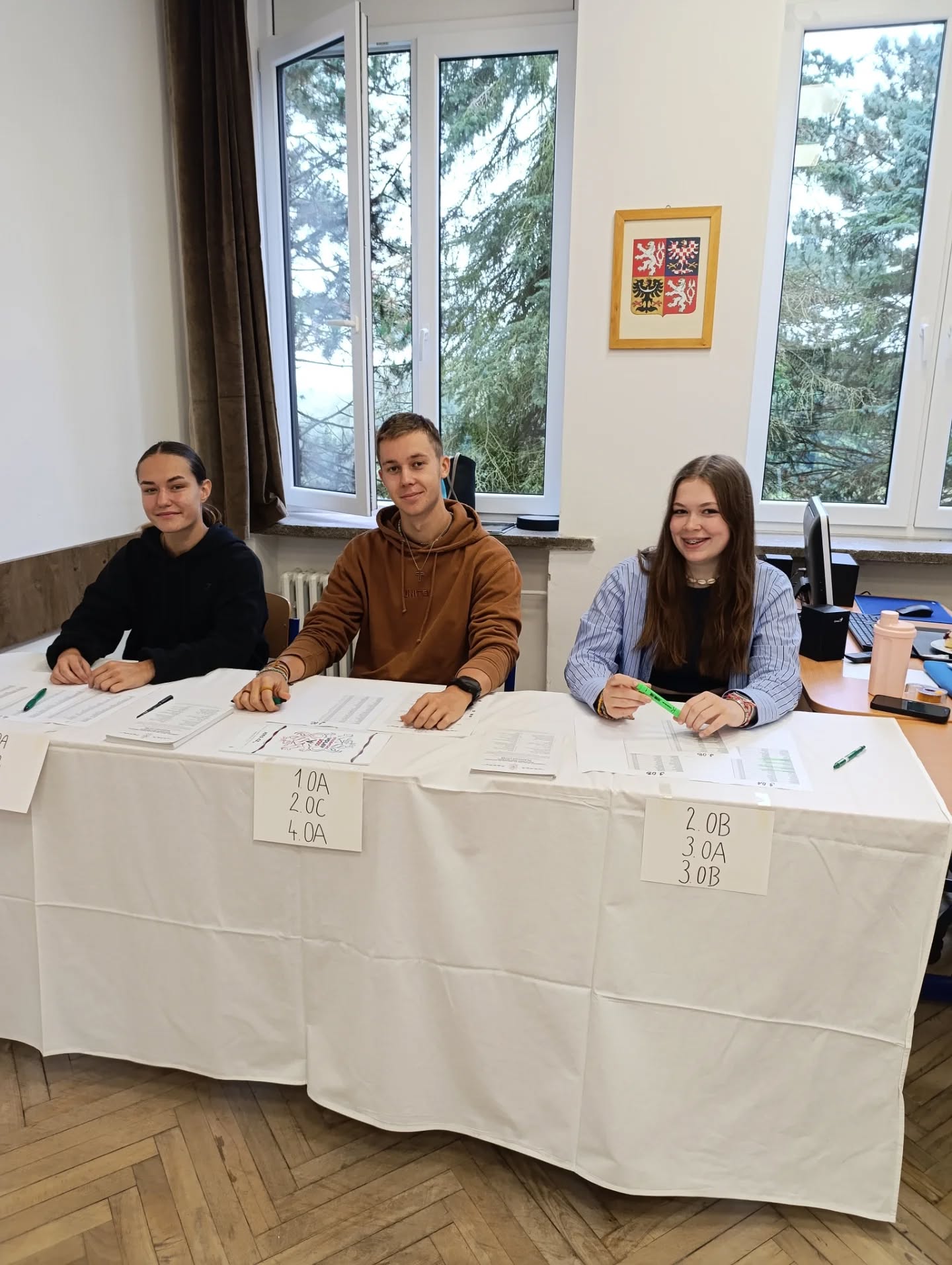 Studenti sedí za stoly s cedulkami tříd a připravují organizaci studentských voleb.