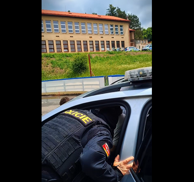 Policista v taktické vestě s nápisem Policie zasahuje u vozidla před budovou školy.
