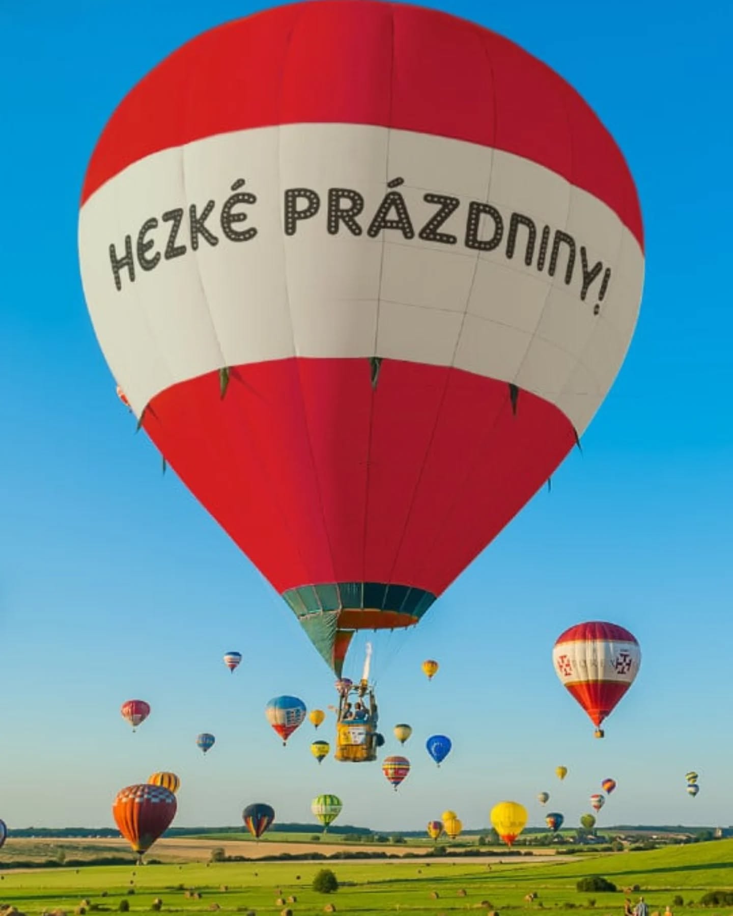 Horkovzdušný balón s nápisem „Hezké prázdniny!“ stoupá nad krajinou plnou dalších balónů.
